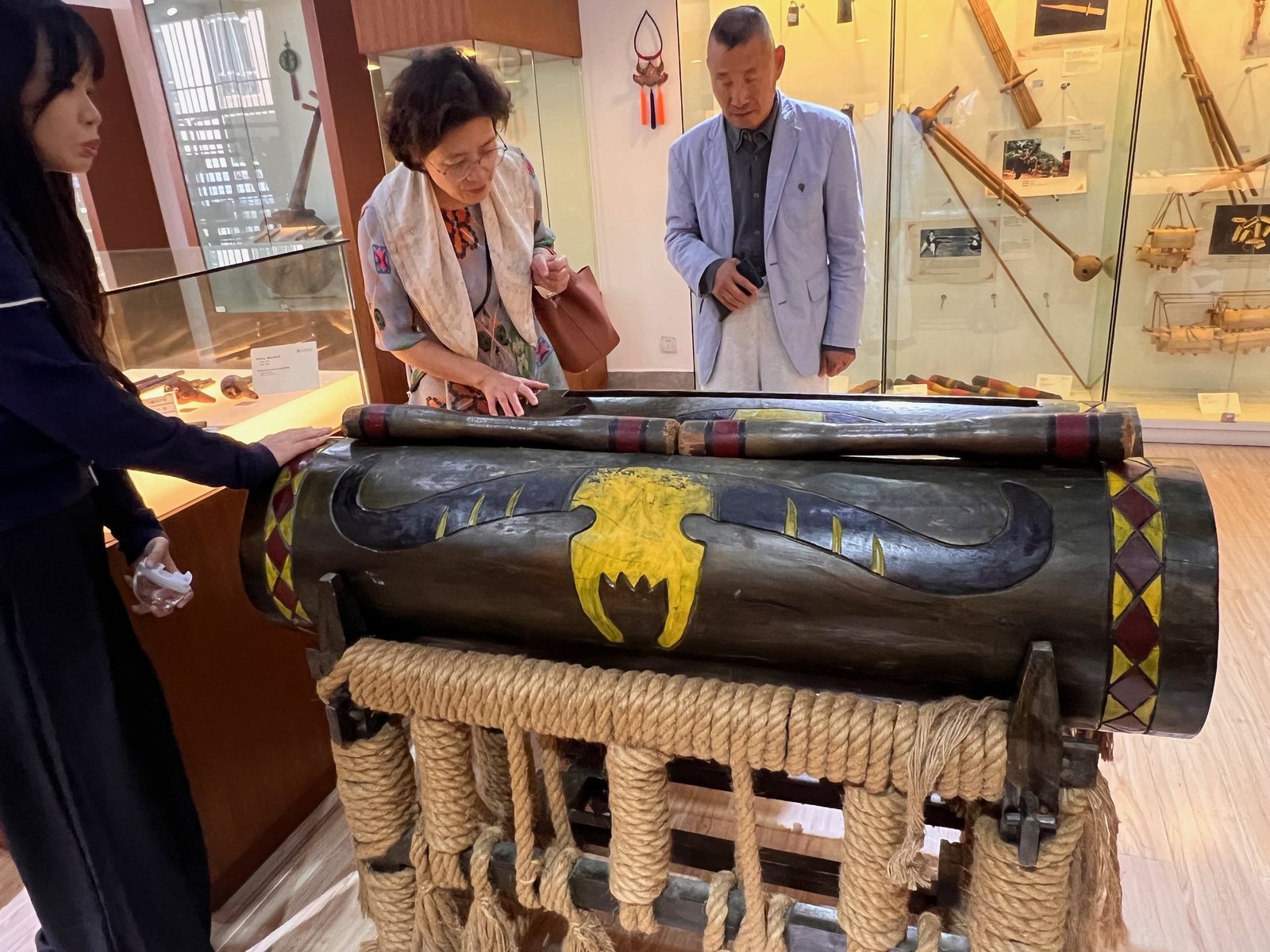 Trois visiteurs examinent un instrument en bois décoré dans une exposition.